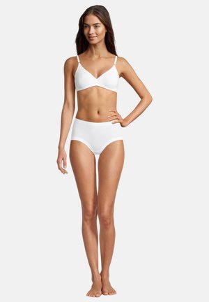 Donna in piedi a piedi nudi, con indosso un reggiseno bianco e mutande a vita alta, una mano sull'anca, su uno sfondo chiaro e semplice.