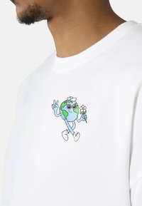 Valge puuvillane T-särk, millel on joonistatud koomiline Maa karakter, kellel on käed, sinised ja rohelised mandrid, suured silmad, kes hoiab lille ja kannab valgeid jalanõusid.