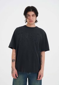 Czarna oversize'owa koszulka z krótkim rękawem, na której widnieje napis "ED HARDY" w ciemnym druku. Posiada luźny krój. Noszona z niebieskimi poszarpanymi dżinsami.
