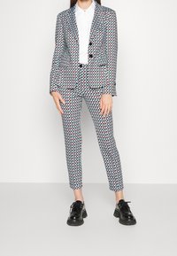 Costume imprimé géométrique coloré en tissu mélangé blanc, bleu clair, rouge et noir. Présente deux boutons sur la veste, un pantalon slim-fit et des chaussures noires.