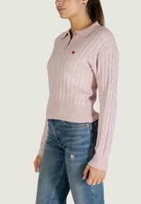 Maglione di colore rosa chiaro a trecce con colletto. Presenta un piccolo emblema a forma di cuore rosso sul petto e polsini e orlo a coste.