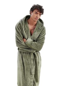Green plush robe with a hood, long sleeves, a tied belt at the waist, and a soft texture. Model stands with arms crossed, displaying the garment.