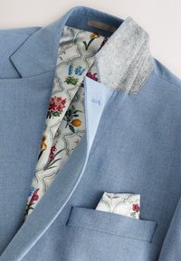 Blazer azzurro chiaro con colletto in feltro grigio. Fodera e fazzoletto da taschino a motivo floreale con fiori colorati su sfondo bianco.