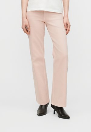 Ženska, oblečena v svetlo rožnate hlače z ravnimi hlačnicami in črne škornje z ostrim sprednjim delom in peto, stoji pred enobarvnim belem ozadju.