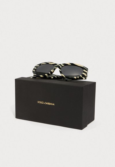 Schwarz-weiße gestreifte Sonnenbrille mit quadratischem Rahmen auf einer schwarzen Box, die ein geprägtes goldfarbenes Logo auf der Vorderseite hat.