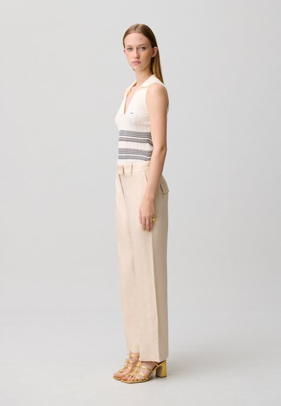 Femme debout de profil portant un haut blanc sans manches à rayures horizontales, un pantalon large beige et des sandales à talons jaunes.