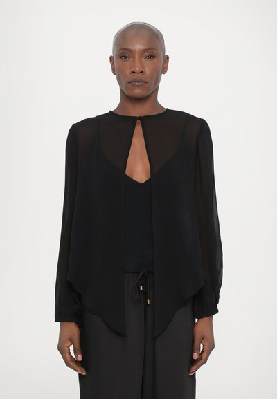 Durchscheinende schwarze Bluse mit einer Schlüssellochöffnung, langen Ärmeln und einem fließenden Design. Getragen über einem schwarzen Oberteil und kombiniert mit schwarzen Hosen.