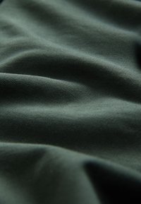 Donkergroen, zacht stof met een gladde textuur. Bevat zachte plooien en draperingen, die subtiele variaties in licht en schaduw tonen.