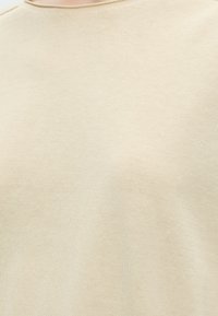 Gros plan d’un tissu tricoté beige clair sur un vêtement à col rond, montrant une texture douce et des détails de couture fins.