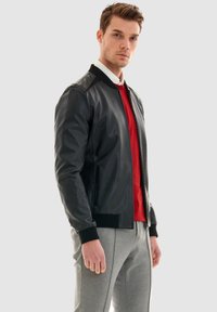 Schwarze Leder-Bomberjacke mit gerippten Bündchen und Kragen. Verfügt über Reißverschlusstaschen und eine taillierte Silhouette. Trägt man über einem roten Pullover.