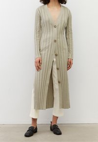 Cardigan lungo a coste di un beige chiaro con scollo a V profondo, dotato di sette bottoni. Indossato sopra pantaloni bianchi e mocassini neri. Texture liscia.