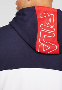 Sudadera marina con acento rojo en la parte posterior, que presenta un diseño de logo en blanco. Confeccionada en suave algodón con textura lisa y diseño a bloques de color.