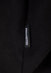 Svart stoffmerke med ordet "FORBUDT" i hvitt, sydd på et glatt svart klesmateriale med en enkel tekstur.