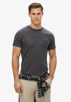 Camiseta gris con un sutil estampado de logo, combinada con pantalones cortos beige claros y una camisa de franela a cuadros verde y rojo atada a la cintura.