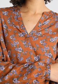 Bruine bloemenblouse met paarse en groene patronen. Heeft een V-hals, plooidetails en lange, gathered mouwen. Zachte stoftextuur.