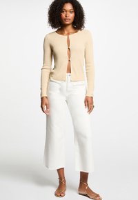 Beige ribbattune cardigan med djup frontöppning och tryckknappar. Matchad med vita vida jeans och gyllene sandaler med banden.