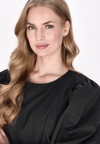 Sort rundhalset top med puffede ærmer, blød tekstur og en let glans. Modellen har langt bølget blond hår og neutral makeup.