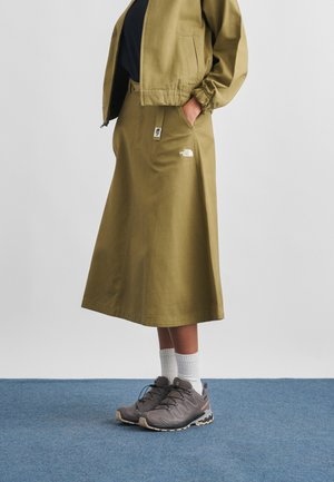 Inimene, kes kannab oliivirohelist North Face seelikut, sobivat jopet, valgeid sokke ja halli matkasussi, seisab sinisel vaibal.