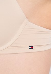 Lähedalt tehtud pilt beežist rinnahoidjast, mille küljes on nähtav õmblus ja väike punase, valge ja sinise värviga ristkülikuline logo, mis on kanga peal nahaga kontaktis.