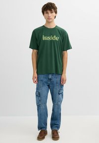 Camiseta de algodón verde con la palabra "inside" impresa en verde claro. Combinada con unos vaqueros cargo azules y unos zapatos marrones de cordones. Diseño simple.