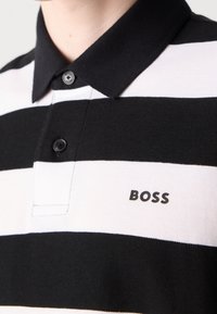Gros plan d'une personne portant un polo rayé horizontal noir et blanc avec un col et des boutons noirs, avec le texte "BOSS" sur la poitrine.