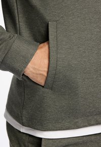 Sudadera verde oliva con tejido texturizado, que cuenta con un bolsillo lateral y un dobladillo blanco contrastante en la parte inferior.