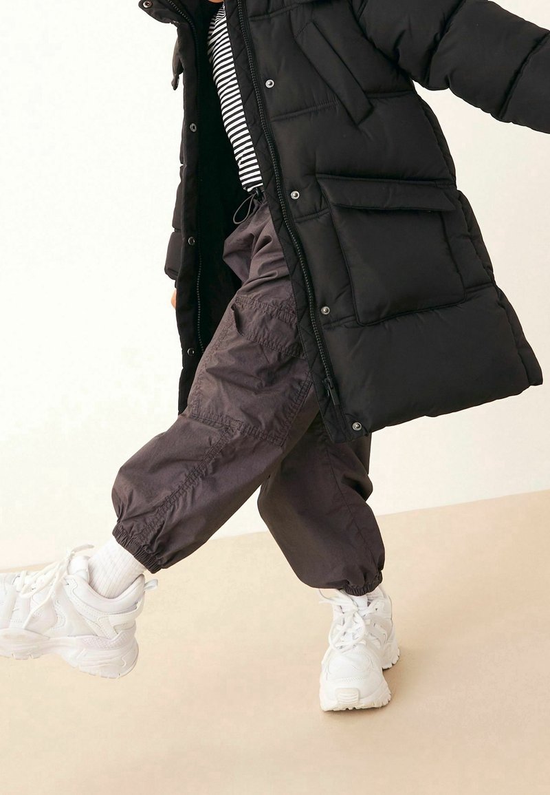 Enfant portant un manteau matelassé noir, une chemise rayée, un pantalon cargo foncé et des baskets blanches, levant une jambe sur un fond uni.