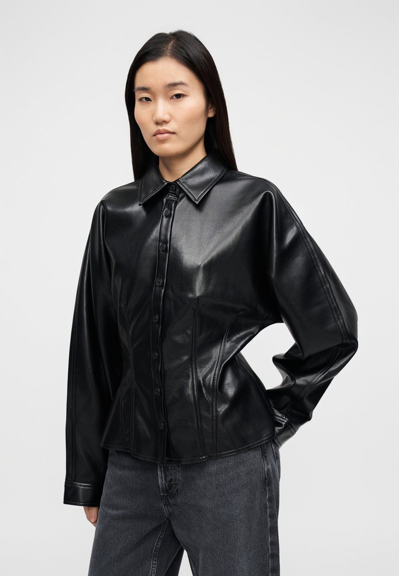 Chemise noire en cuir boutonnée avec une silhouette structurée, un col pointu et des manches longues, présentant des coutures contrastées et une texture brillante.