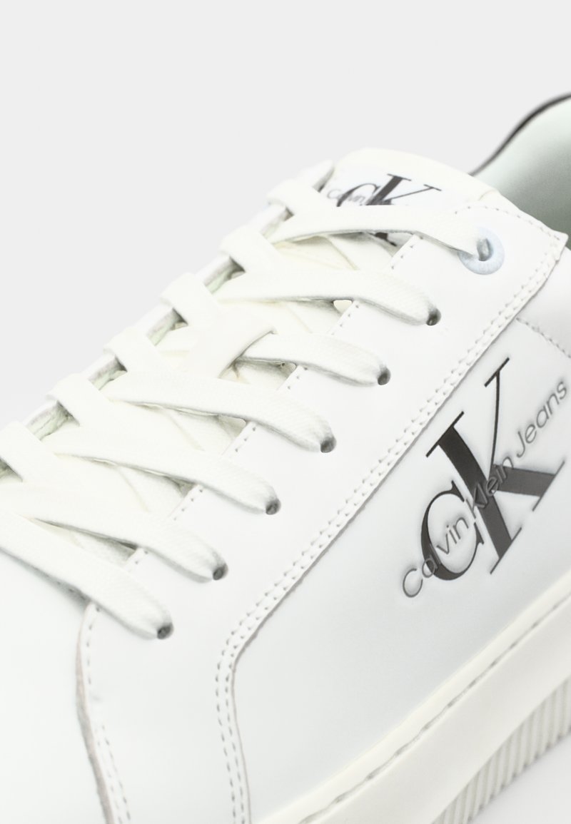 Baskets en cuir blanc avec un design texturé, dotées de lacets plats, d'un logo sur le côté et d'une semelle en caoutchouc avec des lignes horizontales.