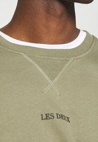 Uomo che indossa una felpa verde chiaro con una maglietta bianca visibile sul colletto, la felpa presenta la scritta "LES DEUX" sul davanti.