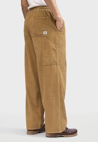 Brown corduroy pants with a high waist, wide-leg design, side pockets, and a small branded patch on the back pocket. Textured fabric.