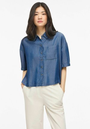 Młoda kobieta o długich ciemnych włosach, ubrana w luźną niebieską koszulę z krótkim rękawem zapinaną na guziki oraz jasne spodnie, z rękami w kieszeniach.