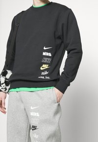 Persona vestida con un sudadera negra y pantalones de chándal grises con múltiples logotipos de Nike, sosteniendo un bolso de patrón en blanco y negro en el hombro.