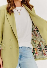Jasnozielona podwójnie zapinana marynarka z kwiatowym wpasowaniem, biała bluzka i jasnoniebieskie dżinsy. Marynarka ma dwa guziki i kieszeń na piersi.
