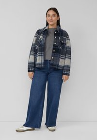 Blau und grau kariertes Flanelljacke mit zwei Brusttaschen, kombiniert mit einem grauen Oberteil und weitem Jeans mit weitem Bein. Weiße und beige Sneaker.