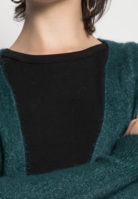 Cardigan fuzzy teal con apertura frontale e maniche lunghe indossato sopra una maglietta nera. Il cardigan ha una texture morbida e una vestibilità rilassata.