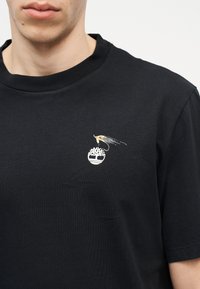 Μαύρο μπλουζάκι με μικρό λευκό λογότυπο δέντρου και πολύχρωμο αγκίστρι ψαρέματος κεντημένο στο αριστερό μέρος του στήθους, φορεμένο από άτομο με ανοιχτόχρωμο δέρμα.