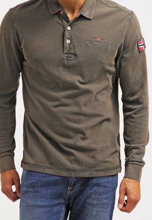 Mężczyzna w oliwkowej, długiej koszulce polo z zapięciem na guziki i naszywką na lewym rękawie, w połączeniu z niebieskimi dżinsami.