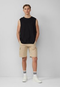 Zwarte mouwloze shirt, beige cargoshorts met zijzakken, witte sneakers en gestreepte sokken; model staande tegen een effen achtergrond.