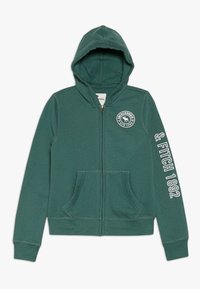 Veste à capuche zippée en mélange de coton vert foncé. Elle comporte deux poches frontales, un logo sur la poitrine et du texte blanc le long de la manche.