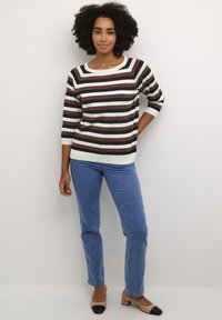 Pull rayé marron et crème avec un décolleté large, manches trois-quarts, associé à un jean bleu clair et des chaussures à talons blocs bicolores.