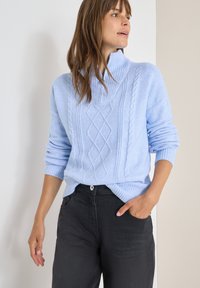 Hellblauer Strickpullover mit hohem Kragen, der über Kabelstrickmuster und gerippte Bündchen verfügt. Kombiniert mit schwarzen Hosen.