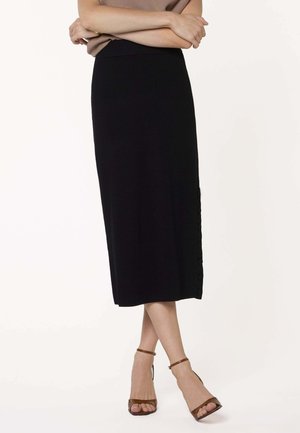 Vrouw draagt een zwarte midirok met knopen aan de zijkant, gecombineerd met bruine enkellaarsjes met enkelbandje, armen gekruist over een beige top.