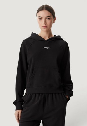 Suorat ruskeat hiukset omaava nainen pukeutuu mustaan Tommy Jeans -huppariin ja yhteensopiviin housuihin, seisten vaaleaa yksiväristä taustaa vasten.