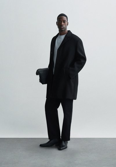 Fekete, gyapjúszerű kabát laza szabással, alatta szürke pulóver, fekete nadrág és hegyes fekete cipő. Egy fekete karcibag az egyik kezében.