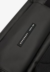 Zwarte rugzak met een gladde, matte textuur. Bevat een rechthoekig logo-opdruk met "SUPERDRY" in het wit. Het ontwerp is gestructureerd en stijlvol.