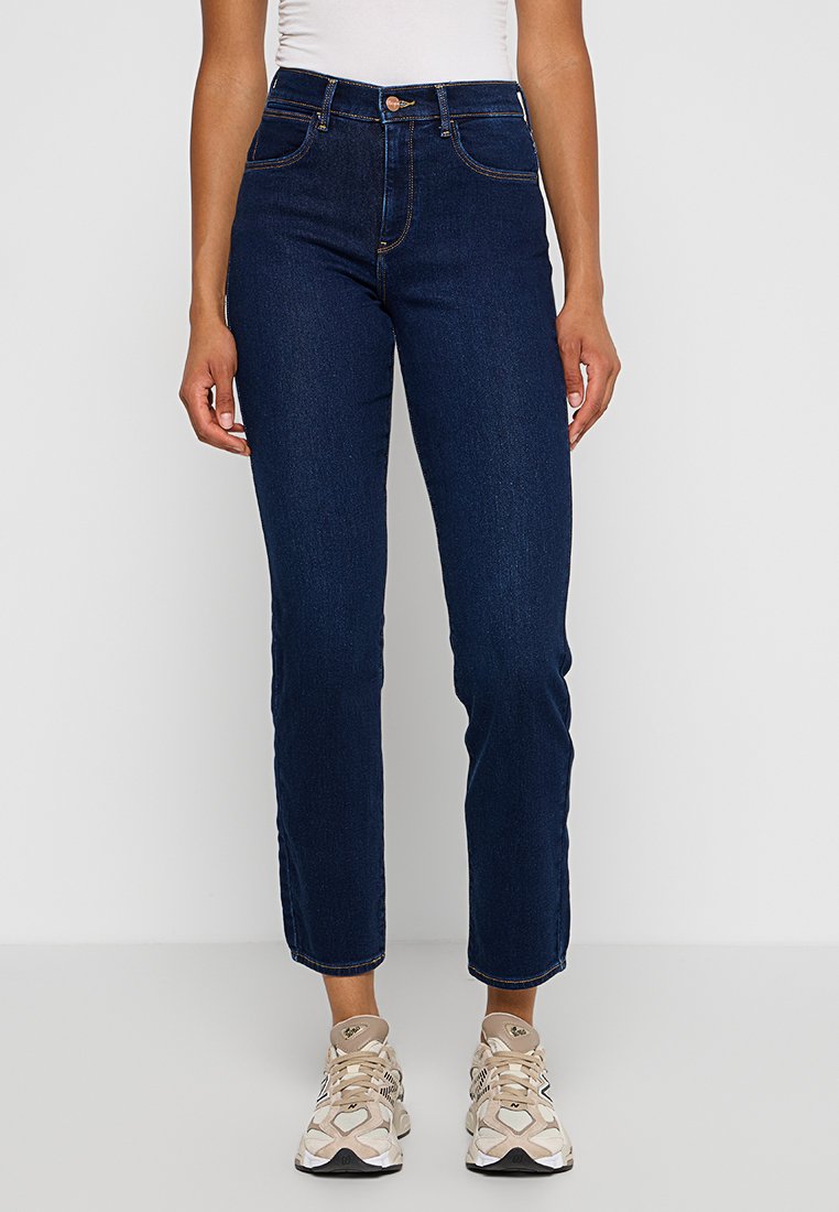 Jean en denim bleu foncé avec une coupe droite, poches avant et détails de bouton en cuivre. Porté avec des baskets beiges.