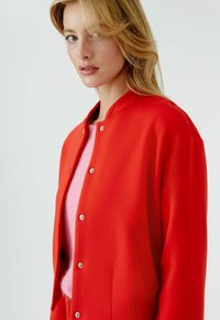 Blonde Frau mit blauen Augen trägt eine leuchtend rote, geknöpfte Jacke über einem weichen rosa Pullover vor einem schlichten, hellen Hintergrund.