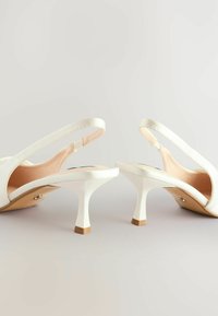 Valkoiset slingback-korkokengät satiinipinnoitteella, avonaisella varpaalla, kapealla kantapäällä ja erottuvalla neliömäisellä kantapääsuojalla. Suora pinta, minimalistinen muotoilu.