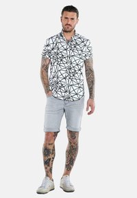 Kurzärmliges schwarzes und weißes Hemd mit geometrischem Muster, hellgraue Shorts und weiße Sneaker. Auffällige Tätowierungen an sichtbaren Armen und Beinen.